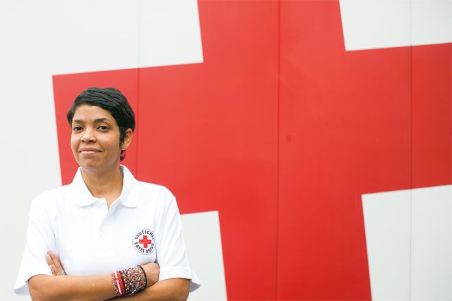 Foto: Eine junge DRK-Mitarbeiterin steht vor einer weißen Wand mit einem großen, roten Kreuz. Sie blickt direkt in die Kamera und wirkt insgesamt zuversichtlich und gelassen.